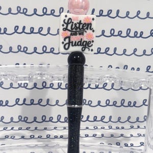 May include: A decorative pen with a black glitter barrel and a silver accent. The pen features a pink bead, a black top, and a white square with the text "We Listen and We Don't Judge" with pink flower accents. The pen is displayed in a clear pen holder.
