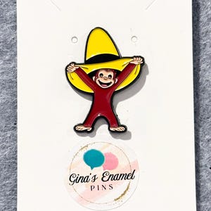 May include: Enamel pin featuring a cartoon monkey wearing a yellow hat. The monkey is red with black accents. The pin is on a white card with the text "Gina's Enamel PINS" in a circular design.