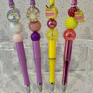 May include: Four colorful pens with different charms attached to the top. The charms include a rainbow with the text "Be Pawsitive", a sun with the text "Radiate Positivity", a honey pot, and a honey pot with a pink and yellow bee.