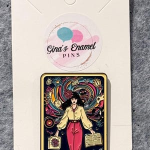 May include: A gold-colored enamel pin featuring a woman with black hair standing in front of a swirling background of colorful abstract shapes and objects. The woman is wearing a yellow shirt, red pants, and holding a book and a folder. The text "THE ADHD" is printed at the bottom of the pin.