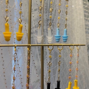 May include: A display of various silicone earplugs with daisy chain earrings. The earplugs are in yellow, blue, white, pink, and black. The earrings are gold with small white flowers.
