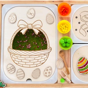 Op de afbeelding: Houten sensorische speelset met Pasen als thema, met een mand gevuld met groen mos, decoratieve eieren en een konijnenontwerp. Inclusief kleurrijke pompons, houten lepels en wortelvormige uitsteekvormen. Ontworpen voor kinderspel.