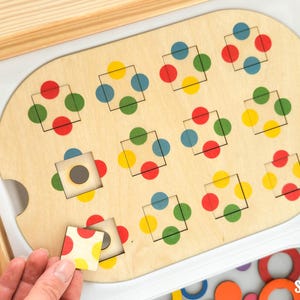 Könnte beinhalten: Pädagogisches Holzspielzeug mit hellem Holzfinish. Das Spielzeug hat ein Brett mit quadratischen Aussparungen und bunten Punktmustern. Eine Hand legt einen passenden Stein ein. Das Spielzeug ist zum Lernen und Spielen konzipiert.