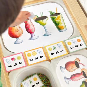 Pode incluir: Um conjunto de atividades educativas para crianças com ilustrações de cocktails coloridos e cartões numéricos. O conjunto inclui imagens impressas de vários cocktails, fatias de fruta, cubos de gelo e números para jogos de contagem e correspondência. A atividade é realizada numa bandeja branca.