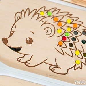 May include: Wooden toy hedgehog with a smiling face and colorful pegs in its quills. The hedgehog is on a light brown wooden base with the text "STUDIO LITTLE EDEN" at the bottom.