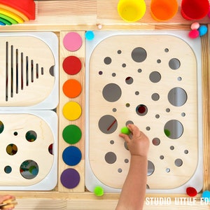 Può includere: Un centro di attività in legno con varie forme e colori. Presenta un arcobaleno, un pannello con un motivo di linee, un pannello con un motivo di cerchi e un pannello con un motivo di cerchi piccoli e grandi. Una mano di bambino è mostrata mentre posiziona una palla verde in uno dei fori circolari.