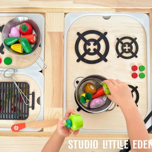 Könnte beinhalten: Ein hölzernes Spielzeug-Küchenset mit einem Herd, einem Grill und einem Topf gefüllt mit bunten Plastikgemüse. Der Herd hat zwei schwarze Brenner und rote und grüne Knöpfe. Der Grill hat einen schwarzen Metallrost. Der Topf ist silberfarben und hat einen Griff. Das Gemüse ist rot, orange, grün und rosa.