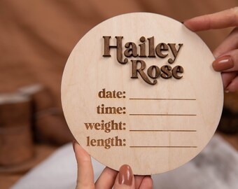 May include: A round wooden plaque with the name "Hailey Rose" engraved in a retro font. The plaque has spaces to record the date, time, weight, and length of a newborn.