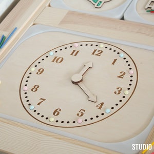 Op de afbeelding: Een houten klok met kleurrijke pinnen rond de rand. De klok heeft de cijfers 1 tot en met 12 en de wijzers staan op 16:00 uur. De klok is onderdeel van een houten speelset.
