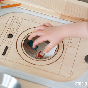 Può includere: Una lavatrice giocattolo di legno con un quadrante bianco e un'apertura rotonda. Una mano di bambino sta inserendo un pezzo di stoffa colorato nell'apertura.