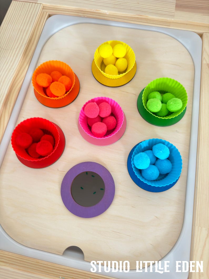 May include: A wooden tray with seven colorful silicone bowls filled with pom poms. The bowls are arranged in a circle around a purple circle with a black center. The text "STUDIO LITTLE EDEN" is printed on the bottom of the tray.
