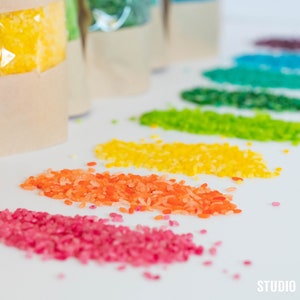 Peut inclure: Riz de couleur arc-en-ciel dans des sachets individuels, avec une petite quantité de chaque couleur répandue sur une surface blanche. Les couleurs sont le rouge, l'orange, le jaune, le vert, le bleu et le violet.