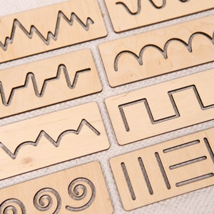 May include: A collection of wooden tracing boards with various cut-out line patterns. The boards are rectangular and made of light-colored wood. The patterns include zigzags, waves, and geometric shapes, designed for tracing and fine motor skill development.