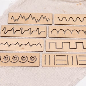 May include: A set of eight wooden tracing boards with various line patterns. The boards are rectangular and feature cut-out designs of wavy lines, zigzags, spirals, and other shapes. The natural wood color provides a neutral background for the patterns.