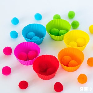 Può includere: Un'attività di ordinamento dei colori arcobaleno con pompon colorati e pirottini per cupcake in silicone. I pompon sono ordinati per colore nei pirottini per cupcake.