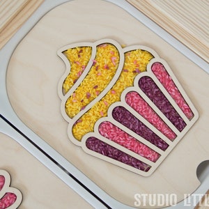 Peut inclure: Un plateau en bois avec une forme de cupcake découpée. Le cupcake est rempli de riz jaune, rose et violet. Le plateau est étiqueté "STUDIO LITTLE EDEN".