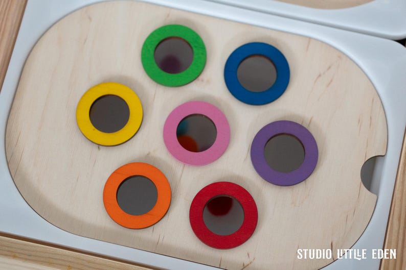 May include: A wooden toy with seven colorful circles, each with a hole in the center. The circles are yellow, green, blue, pink, purple, orange, and red. The toy is designed for children to drop balls through the holes.