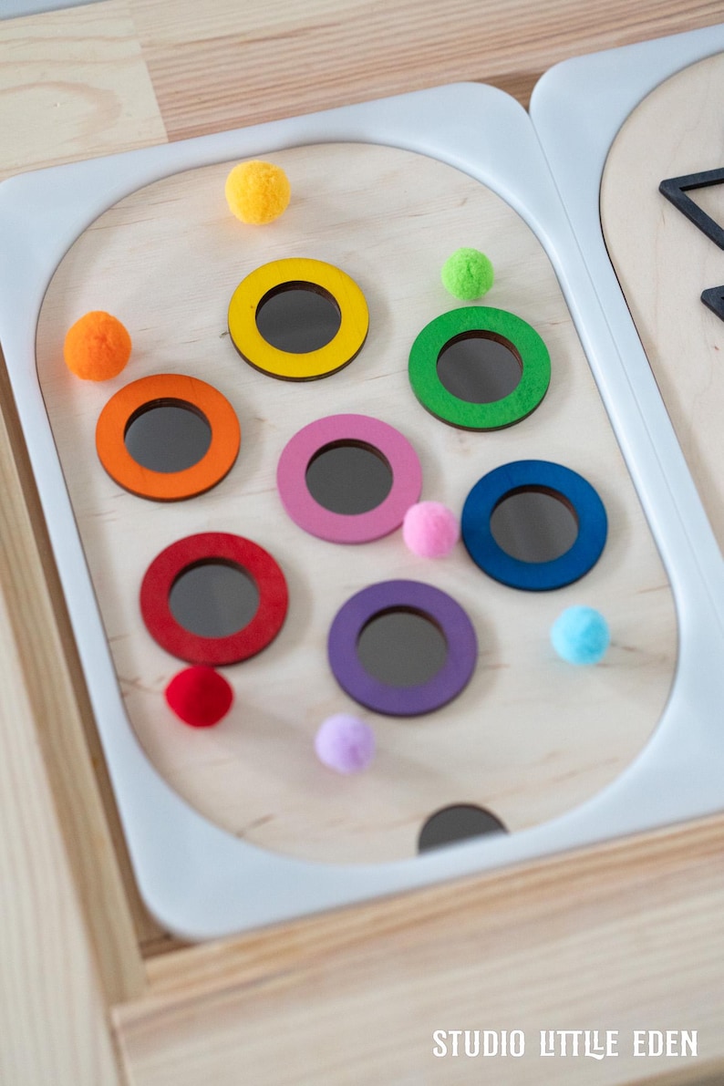 May include: A wooden toy with colorful rings and pom-poms. The rings are yellow, orange, red, pink, purple, blue, and green. The pom-poms are yellow, orange, red, pink, purple, blue, and green.