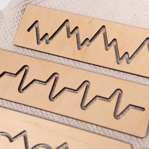 May include: Close-up of several light-colored wooden rectangles with various wave-like patterns cut out. The shapes resemble sound waves or heart rate monitors. The wood has a natural, unfinished appearance, and the background is a textured, off-white fabric.