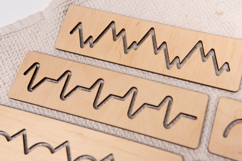 May include: Close-up of several light-colored wooden rectangles with various wave-like patterns cut out. The shapes resemble sound waves or heart rate monitors. The wood has a natural, unfinished appearance, and the background is a textured, off-white fabric.