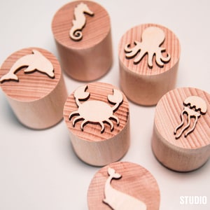 May include: Six wooden stamps with different sea creatures carved into them. The stamps are in the shape of a circle and have a natural wood finish. The sea creatures include a dolphin, a seahorse, an octopus, a crab, a jellyfish, and a whale.