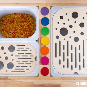 Könnte beinhalten: Ein hölzerner Sensoriktisch mit zwei weißen Kunststoffbehältern, von denen einer mit orangefarbenen und gelben Nudeln gefüllt ist, sowie zwei hellen Holzabdeckungen mit kreisförmigen und schlitzartigen Ausschnitten. Ein Mittelsteg aus Holz enthält sieben bunte Holzscheiben.