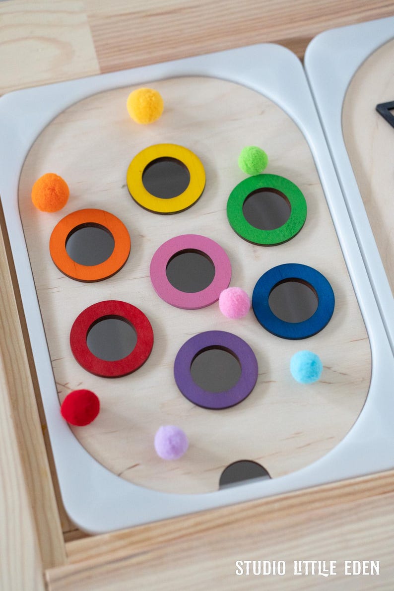 May include: A wooden toy with a white frame and colorful rings. The rings are yellow, green, orange, pink, red, purple, and blue. There are small pom-poms in various colors scattered around the rings.