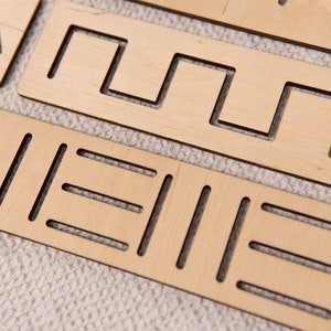 May include: Close-up of several light-colored wooden shape sorter pieces. Each piece has a unique pattern of cut-out shapes, including lines, squares, and curves. The pieces are arranged on a textured, off-white surface.