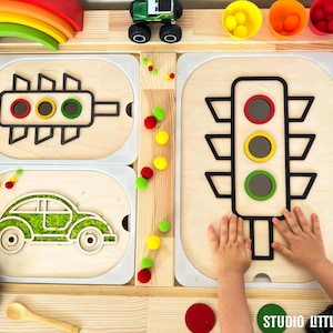 May include: A wooden toy set with a traffic light, car, and steering wheel. The traffic light has red, yellow, and green lights. The car is green and has a white outline. The steering wheel is black and has a white rim. The toy set is on a wooden table with a rainbow arch in the background.