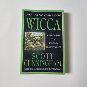 May include: A paperback book titled Wicca: A Guide for the Solitary Practitioner by Scott Cunningham. The black cover features green text, a green border, and a central illustration of a person sitting in a garden. It states Over 400,000 Copies Sold.
