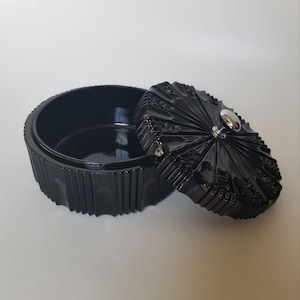 May include: A black, round trinket box with a lid. The box has a ribbed exterior and the lid features a radial design with a silver knob. The box is open, revealing the interior.