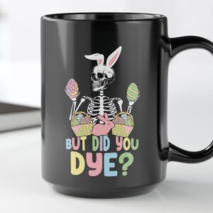 Puede incluir: Taza de café negra con un esqueleto blanco que lleva una diadema de orejas de conejo y sostiene huevos de Pascua. La taza dice "But Did You Dye?"