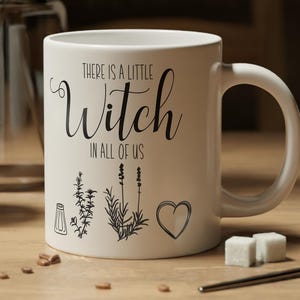 May include: A white ceramic mug featuring the black printed text "There is a little Witch in all of us" along with small line art illustrations of a salt shaker, sprigs of herbs, and a heart symbol on the front surface of the mug.