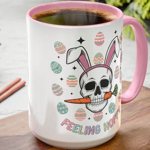 Puede incluir: Taza de café de cerámica blanca con un borde rosa. La taza presenta una calavera de dibujos animados con orejas de conejo rosas y sosteniendo una zanahoria en los dientes. La taza está rodeada de huevos de Pascua coloridos y el texto "Feeling Hoppy".