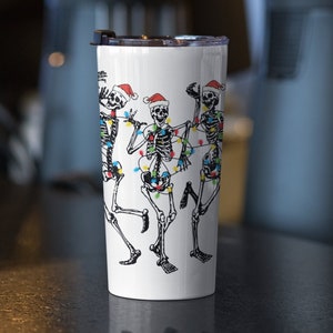 May include: White stainless steel tumbler with a design of three skeletons wearing Santa hats and Christmas lights.