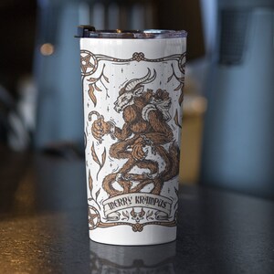 May include: White stainless steel tumbler with a black lid. The tumbler features a brown and white illustration of a mythical creature with the text "Merry Krampus".