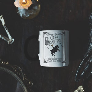 May include: White ceramic mug with a black handle. The mug has a black and white graphic of a person riding a horse with the text "Sleepy Hollow and Dead Breakfast" and "Come and lay your weary head to rest".