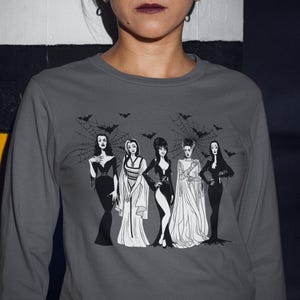 May include: A charcoal gray long-sleeve shirt featuring a black and white graphic print of five iconic horror-themed female characters standing together in front of a spiderweb background with several small bats flying above them.