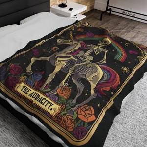 May include: A black and gold tarot card design featuring a skeleton riding a rainbow unicorn. The card is titled "The Audacity."