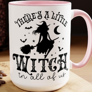 May include: A white ceramic mug with a light pink interior and handle. The front features a black graphic of a witch on a broomstick, stars, bats, and a crescent moon, with the text "THERE'S A LITTLE WITCH in all of us" printed in black.