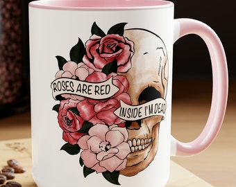 May include: White ceramic mug with a pink rim and handle. The mug features a black and white skull with a floral design. The flowers are pink and red. The text on the mug reads "Roses are red, inside I'm dead."