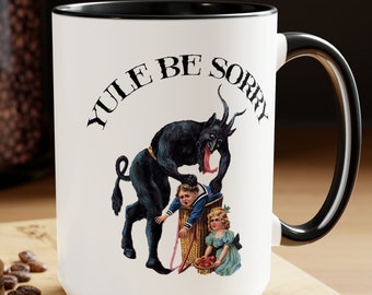 Puede incluir: Taza de cerámica blanca con borde y asa negros. La taza presenta una ilustración en blanco y negro de un diablo con una lengua larga, sosteniendo a un niño en una cesta. El texto "YULE BE SORRY" está impreso en negro en la taza.