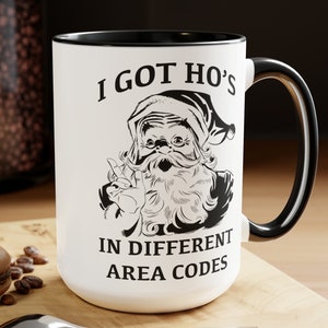 Op de afbeelding: Een witte keramische mok met een zwarte rand en handvat. De mok heeft een zwart-witte illustratie van de kerstman met de tekst "I got ho's in different area codes".