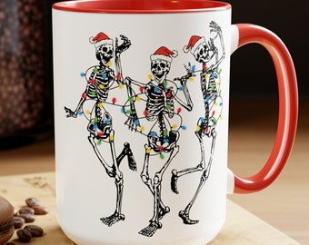 May include: White ceramic mug with a red rim and handle. The mug features a festive design of three skeletons wearing Santa hats and tangled in Christmas lights.