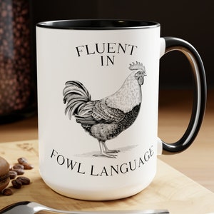 May include: White ceramic mug with a black rim and handle. The mug features a black and white illustration of a rooster with the text "Fluent in Fowl Language".