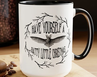 Puede incluir: Taza de cerámica blanca con interior y asa negros. La taza presenta el texto "HAVE YOURSELF A BATTY LITTLE CHRISTMAS" con una ilustración de murciélago, rodeada por una corona de ramas. Una cuchara y un macaron están sobre la superficie de madera.