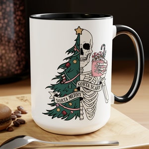 May include: White ceramic mug with a black rim and handle. The mug features a black and white illustration of a skeleton dressed as a Christmas tree. The skeleton is holding a pink mug with a snowflake design. The text "Sorta Merry" and "Sorta Scary" is written on the mug.