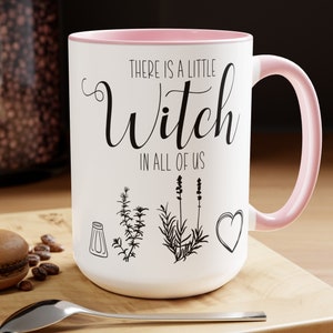 May include: White ceramic mug with a pink rim and handle. The mug has a black graphic that says "There is a little witch in all of us." The graphic also includes a salt shaker, rosemary, thyme, lavender, and a heart.