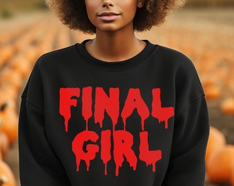 Może przedstawiać: Czarny sweter z czerwonym nadrukiem graficznym, na którym widnieje napis "FINAL GIRL" w kroplących literach. Sweter nosi osoba o kręconych włosach, stojąca na polu z dyniami.