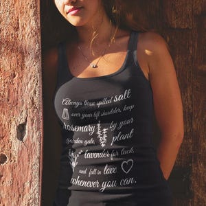May include: A black tank top with white text, paired with denim shorts. The tank top features a botanical-themed quote. The person is leaning against a weathered, orange-toned wall and a wooden door.